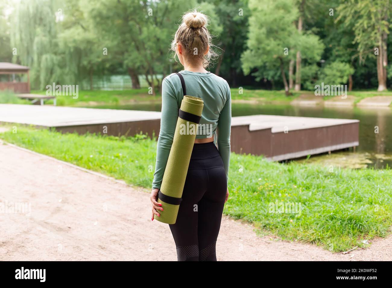 Vista posteriore, una donna snella nel parco in estate, con un tappetino verde da palestra dietro la schiena Foto Stock