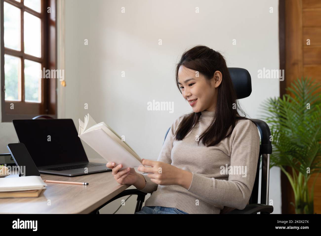 Ritratto della donna asiatica che legge un libro mentre studia l'apprendimento online sul laptop a casa. Foto Stock