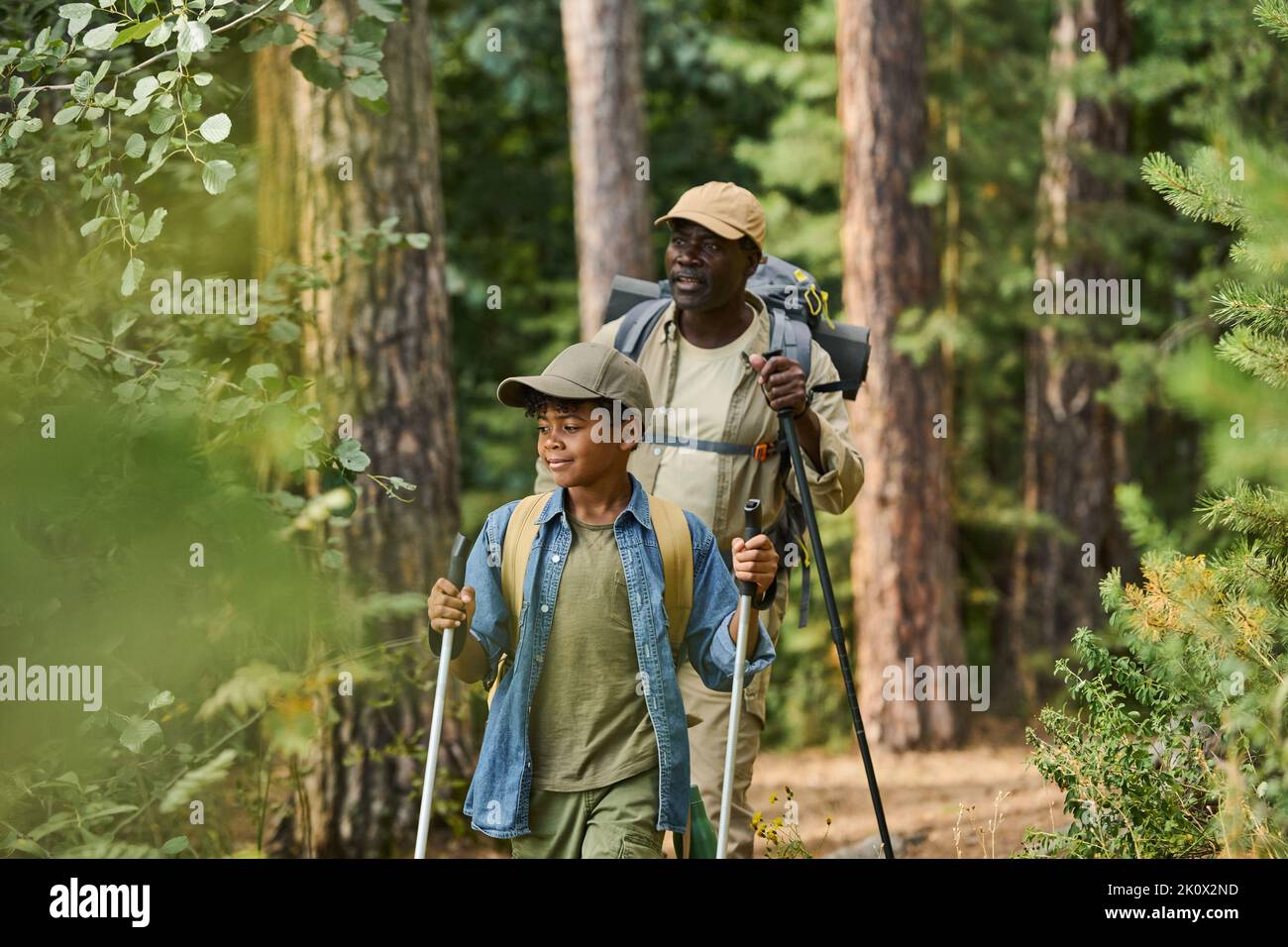 L'uomo nero maturo e suo nipote con bastoni da trekking che si muovono lungo il sentiero lungo gli alberi verdi mentre si gode viaggio zaino Foto Stock