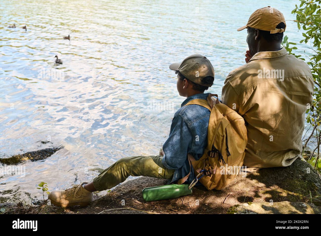 Vista posteriore del giovane ragazzo afro-americano con zaino e suo nonno seduto di fronte al lago o al fiume durante la sosta Foto Stock