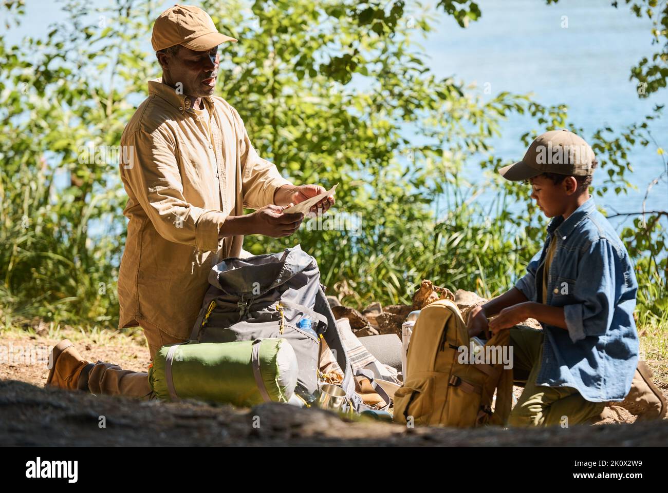 Un anziano uomo afroamericano che prende un piccolo pacchetto con cibo dal suo zaino mentre si trova in piedi sulle ginocchia sul sentiero davanti al nipote Foto Stock