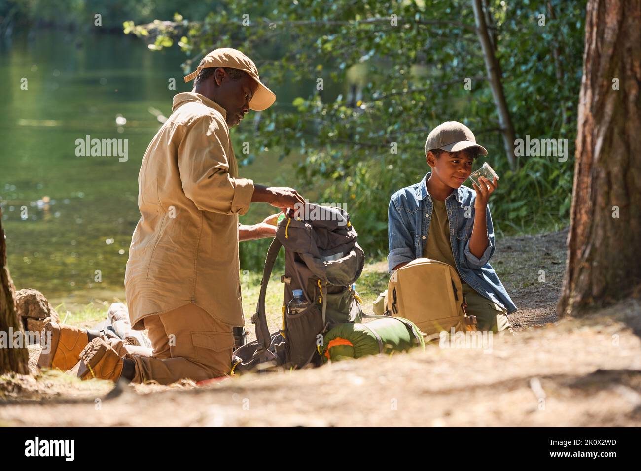 Un anziano uomo afroamericano che disimballa lo zaino mentre si trova in ginocchio sul sentiero davanti al nipote con un vasetto di cavallette Foto Stock