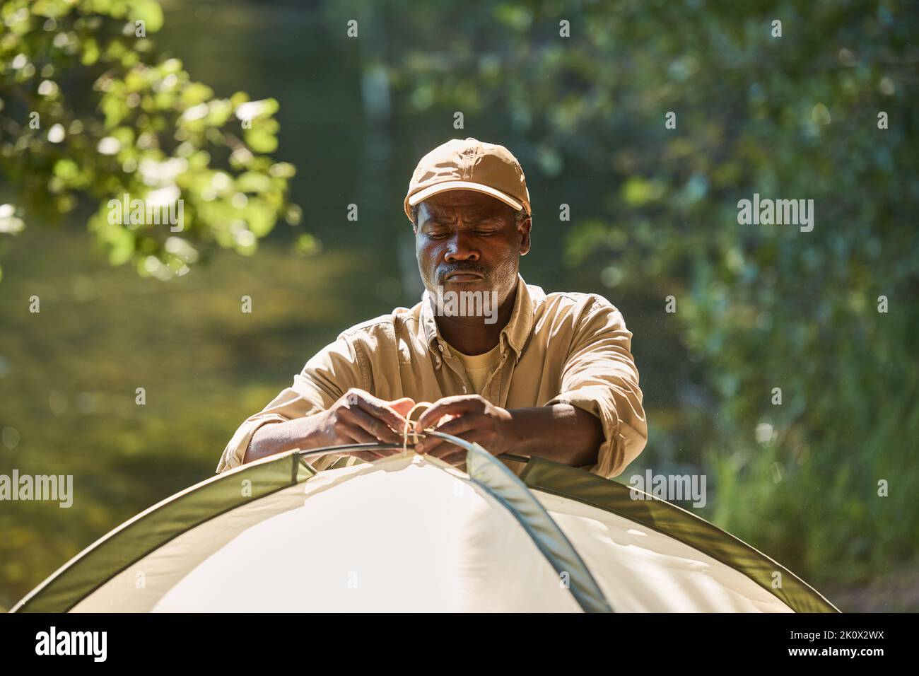 Serio zaino in spalla afroamericano anziano che digita il nodo in cima alla tenda mentre lo mette nella foresta durante il viaggio di escursione nel fine settimana Foto Stock