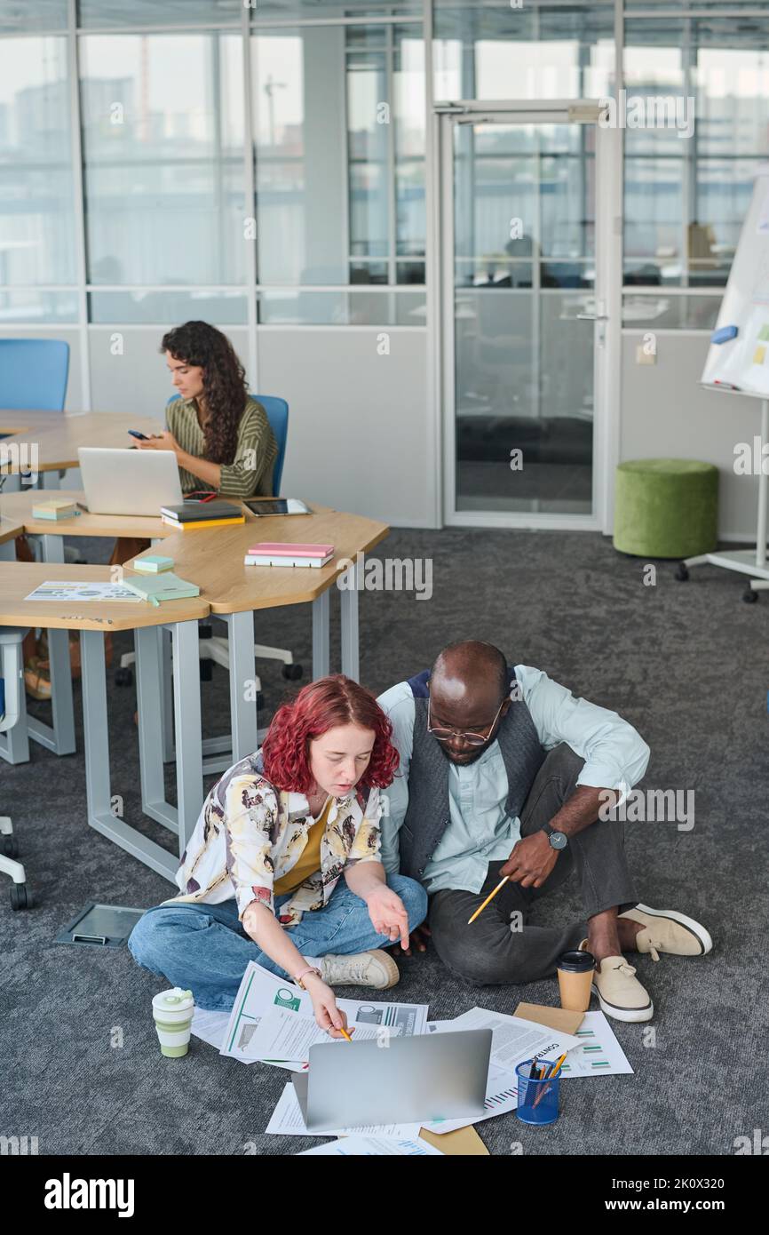 Giovane donna d'affari che indica lo schermo del portatile durante la presentazione di nuovi punti di progetto e discuterli con l'uomo afroamericano Foto Stock