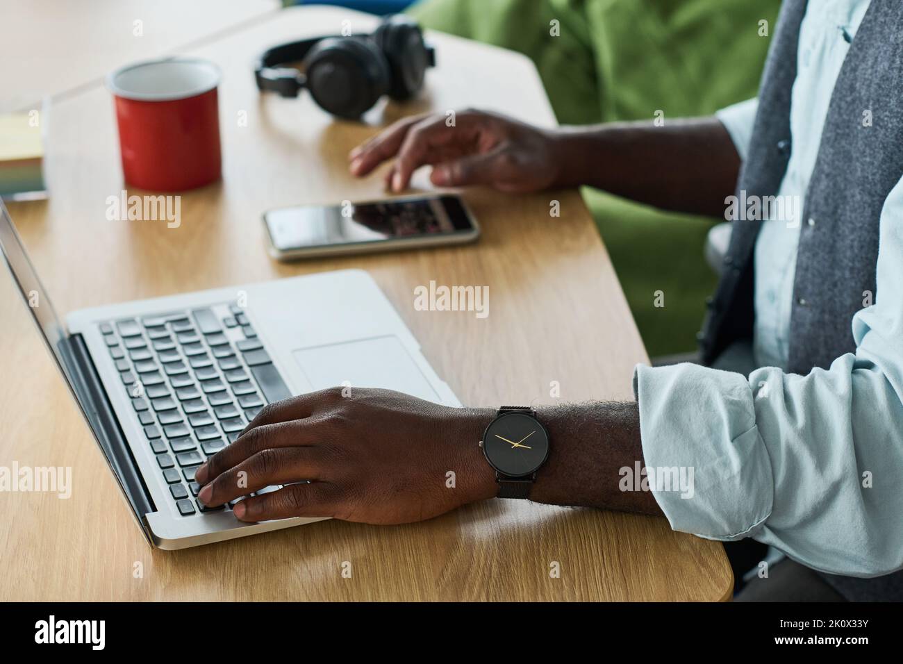 Le mani di un giovane uomo nero in un abbigliamento da ufficio tengono la mano sulla tastiera del computer portatile mentre si scorre sullo smartphone sul posto di lavoro Foto Stock