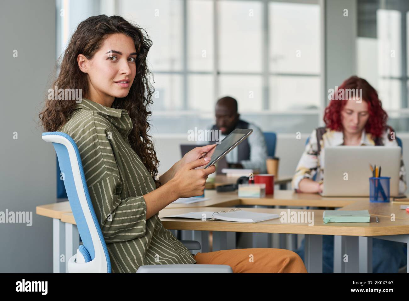 Giovane donna d'affari di successo con un tablet seduto sul posto di lavoro in ufficio tra i suoi colleghi e guardando la fotocamera mentre si lavora in rete Foto Stock