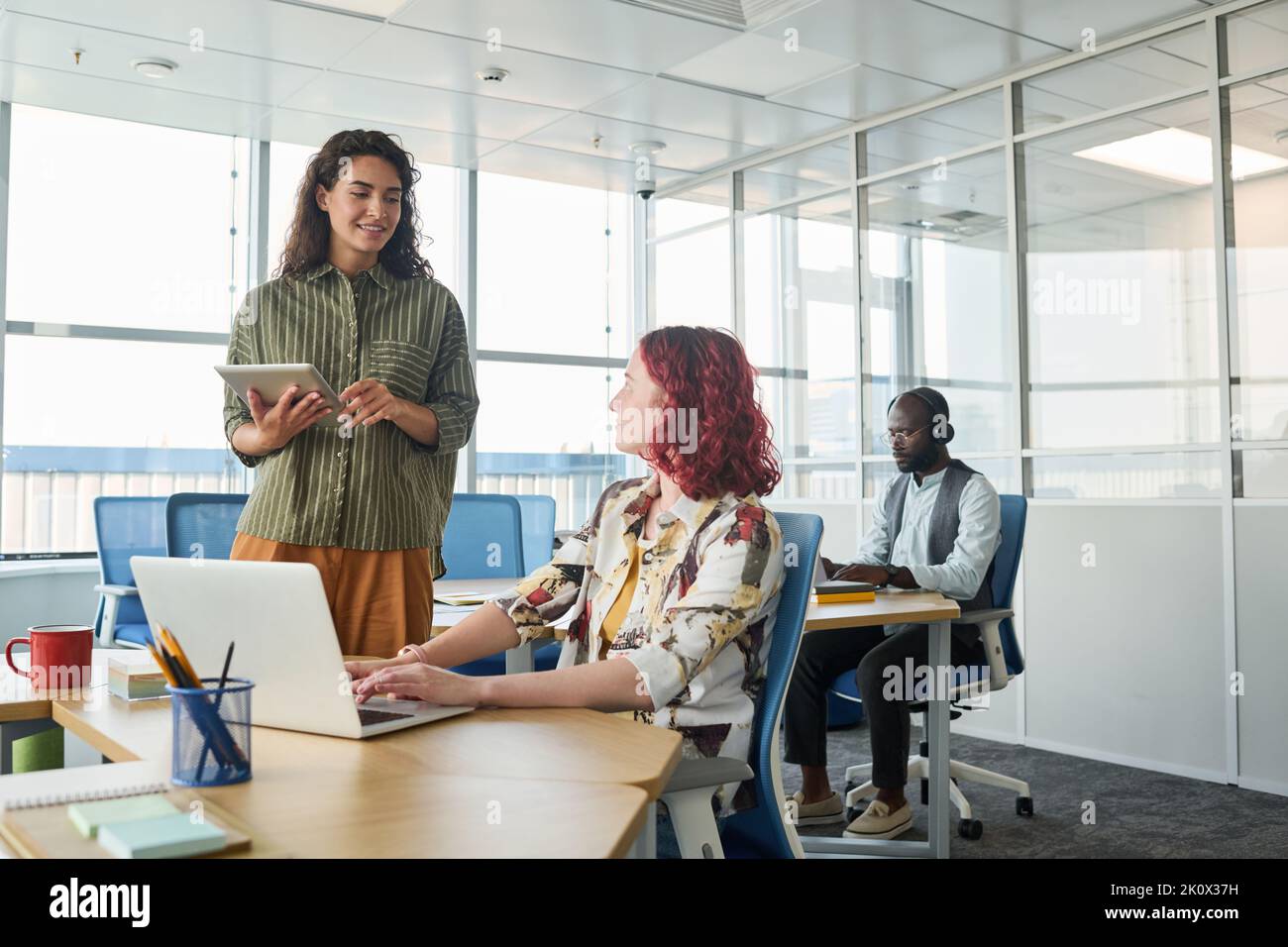 Felice giovane lavoratore con un tablet digitale che comunica con una collega seduto sul posto di lavoro di fronte al notebook durante la conversazione Foto Stock