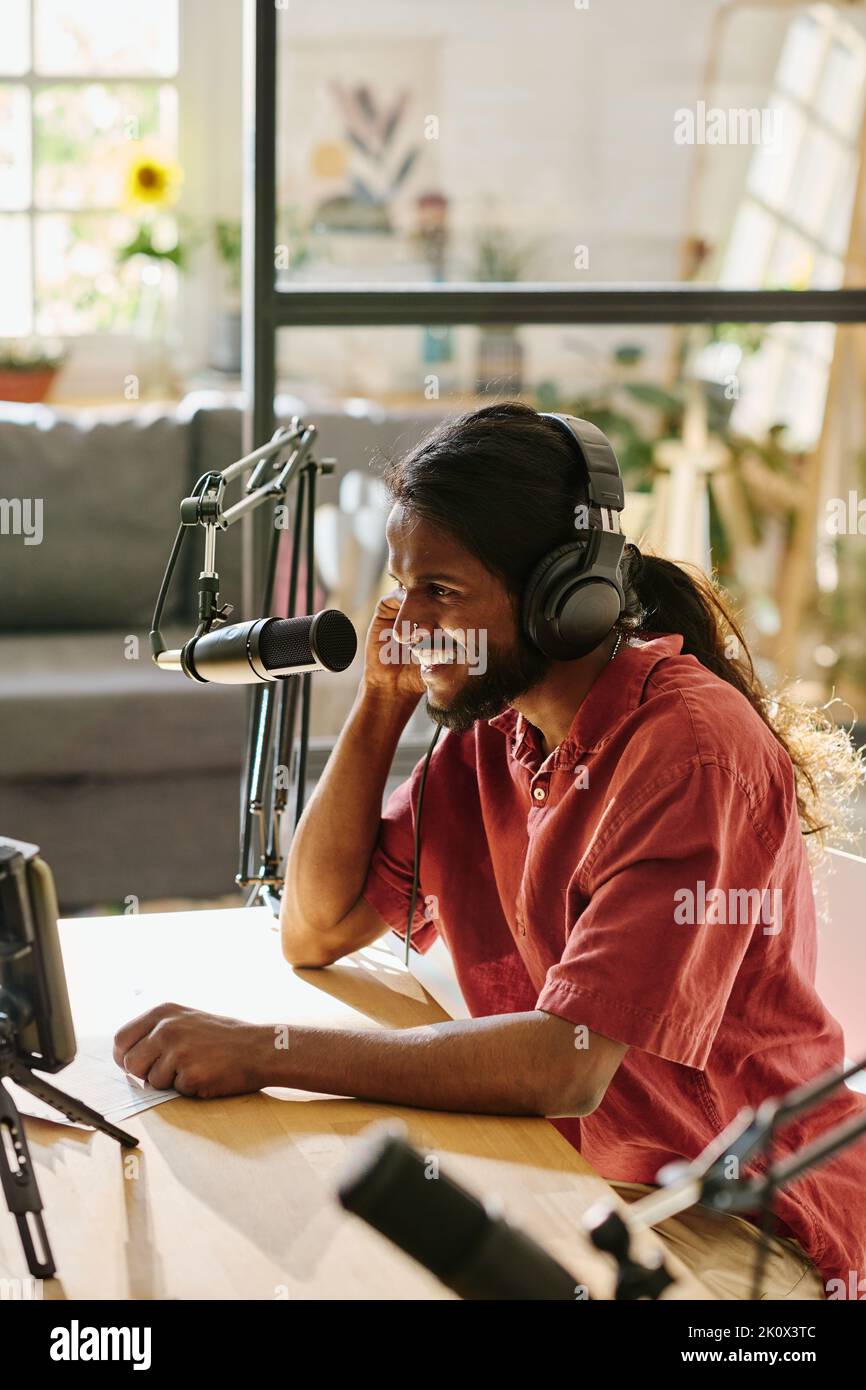 Felice giovane uomo con capelli lunghi scuri che registra nuovi audio o video per il pubblico online dalla scrivania in studio mentre parla nel microfono Foto Stock