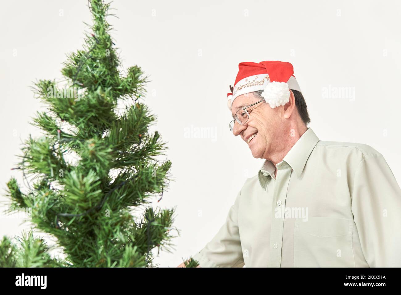 Felice uomo anziano ispanico sorridendo mentre inizia il processo di decorazione del suo albero di Natale indossando un cappello rosso di Babbo Natale con il testo Merry Christ Foto Stock