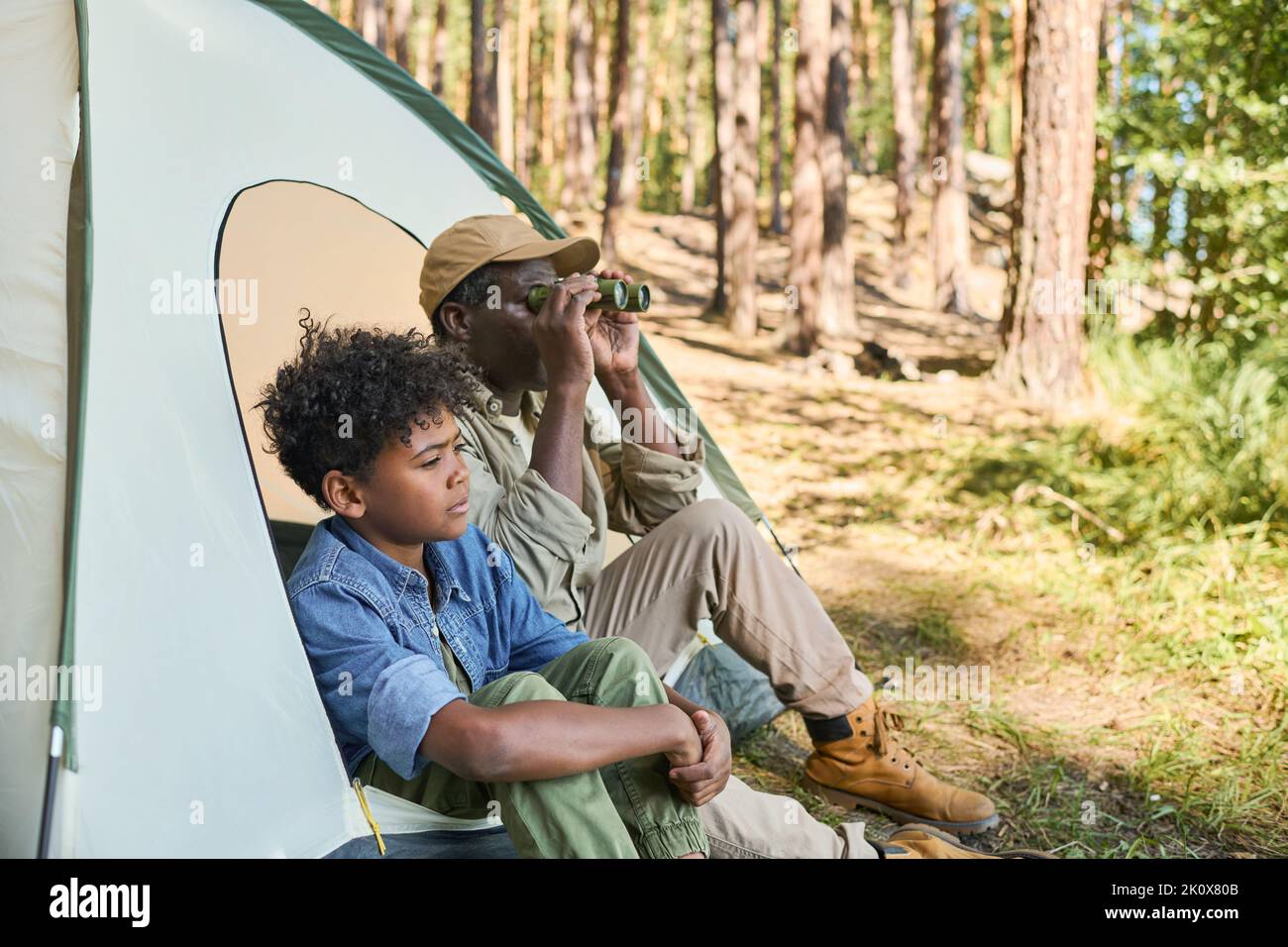 Uomo nero maturo con binocolo per i suoi occhi seduti in tenda accanto al suo nipote in casualwear mentre entrambi riposano durante il viaggio di escursione Foto Stock