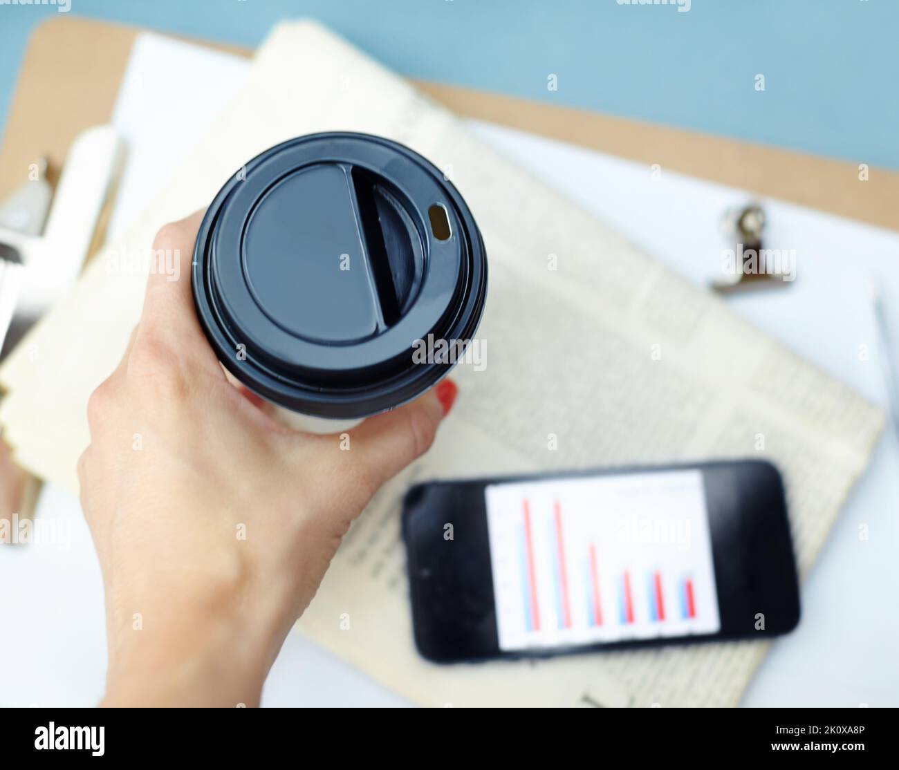 Una tazza di caffè veloce per continuare. Tenere una tazza usa e getta sopra uno smartphone, una clip per raccoglitori, un giornale e una clipboard. Foto Stock