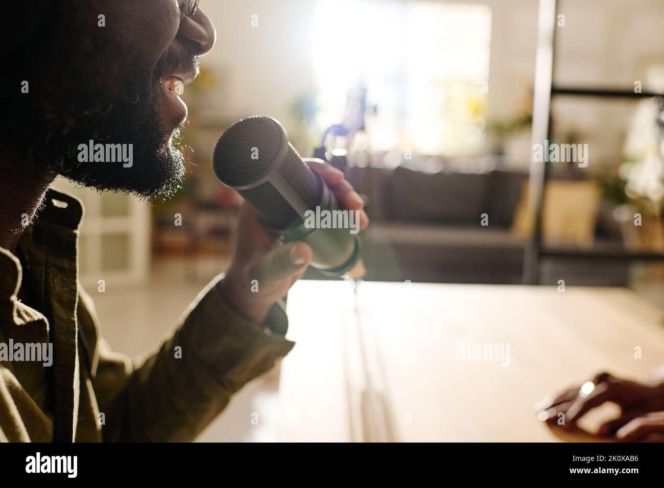 Primo piano di un giovane uomo nero allegro che parla al microfono mentre si siede sul posto di lavoro di fronte alla telecamera e registra file audio Foto Stock