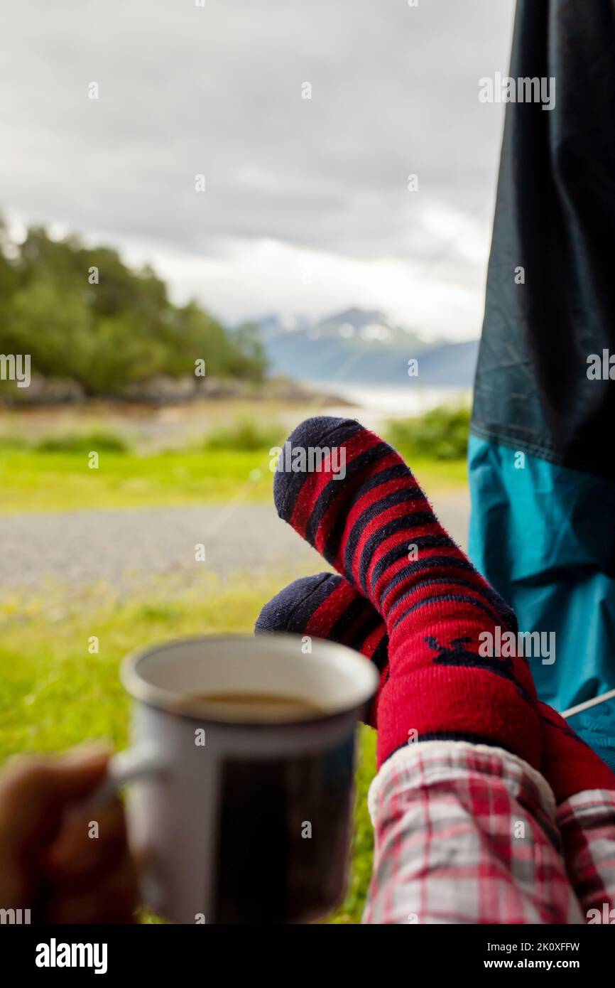 Donna, con calze calde, seduta su una sedia da campeggio di fronte a una tenda, godendo di una tazza di caffè e di una vista incredibile in Norvegia Foto Stock