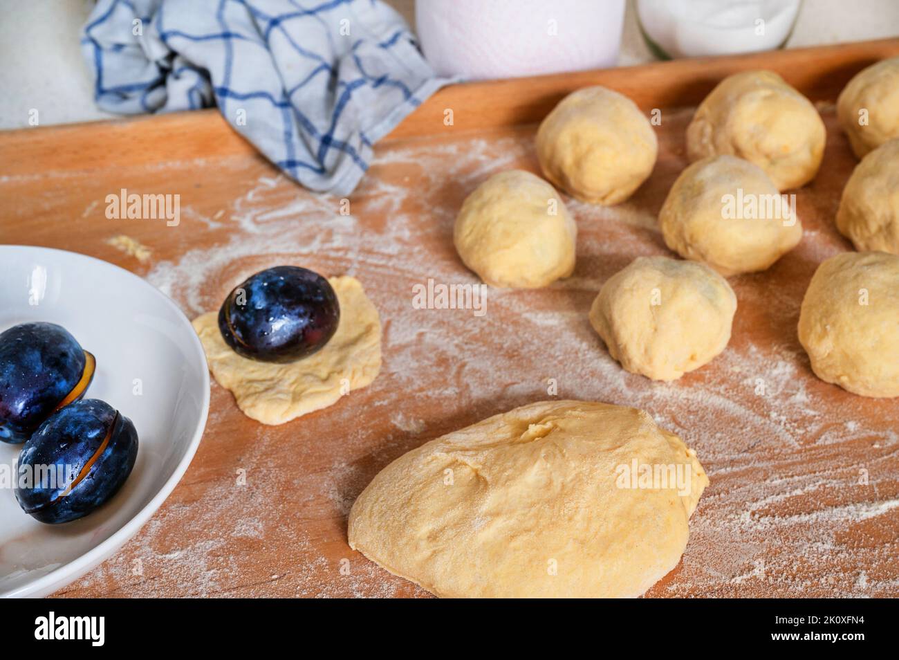 Plum su piatto e su lievito pasta, tovagliolo, bicchiere con zucchero e carta asciugamano da cucina su tavola di legno cosparso di farina, primo piano. Preparazione della prugna dolce Foto Stock