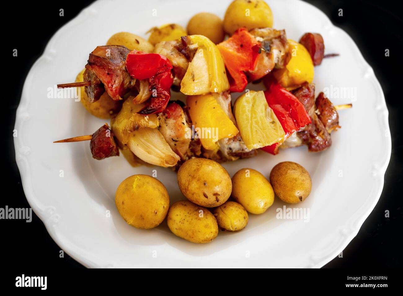 Spiedino con pezzi di carne di maiale, paprika rossa, salsiccia, pancetta e cipolla e patate bollite in pelle su piatto bianco, fondo nero, primo piano. Foto Stock