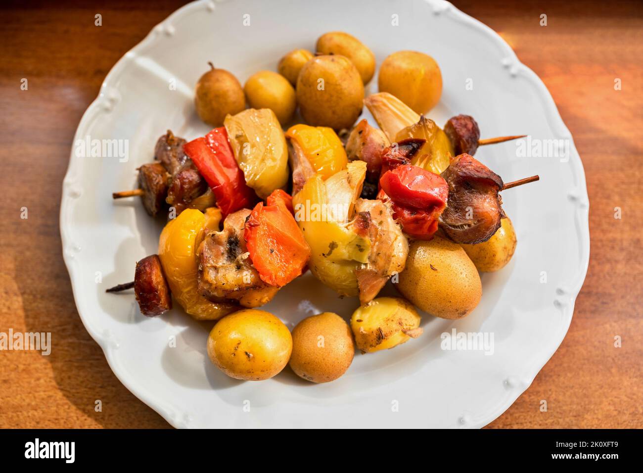 Spiedino con carne di maiale, paprika rossa, salsiccia, pancetta e cipolla e patate bollite in pelle su piatto bianco su tavolo di legno, primo piano. Foto Stock