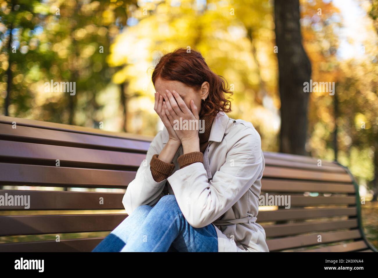 Disappaiata donna caucasica millenaria con capelli rossi in impermeabile siede sulla panca chiude gli occhi con le mani, grida nel parco Foto Stock