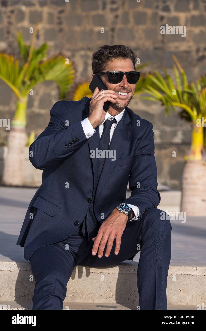 Uomo d'affari felice in tuta e occhiali da sole sorridenti e guardando via mentre si siede sul passo e rispondendo alla chiamata telefonica sulla strada illuminata dal sole della città Foto Stock