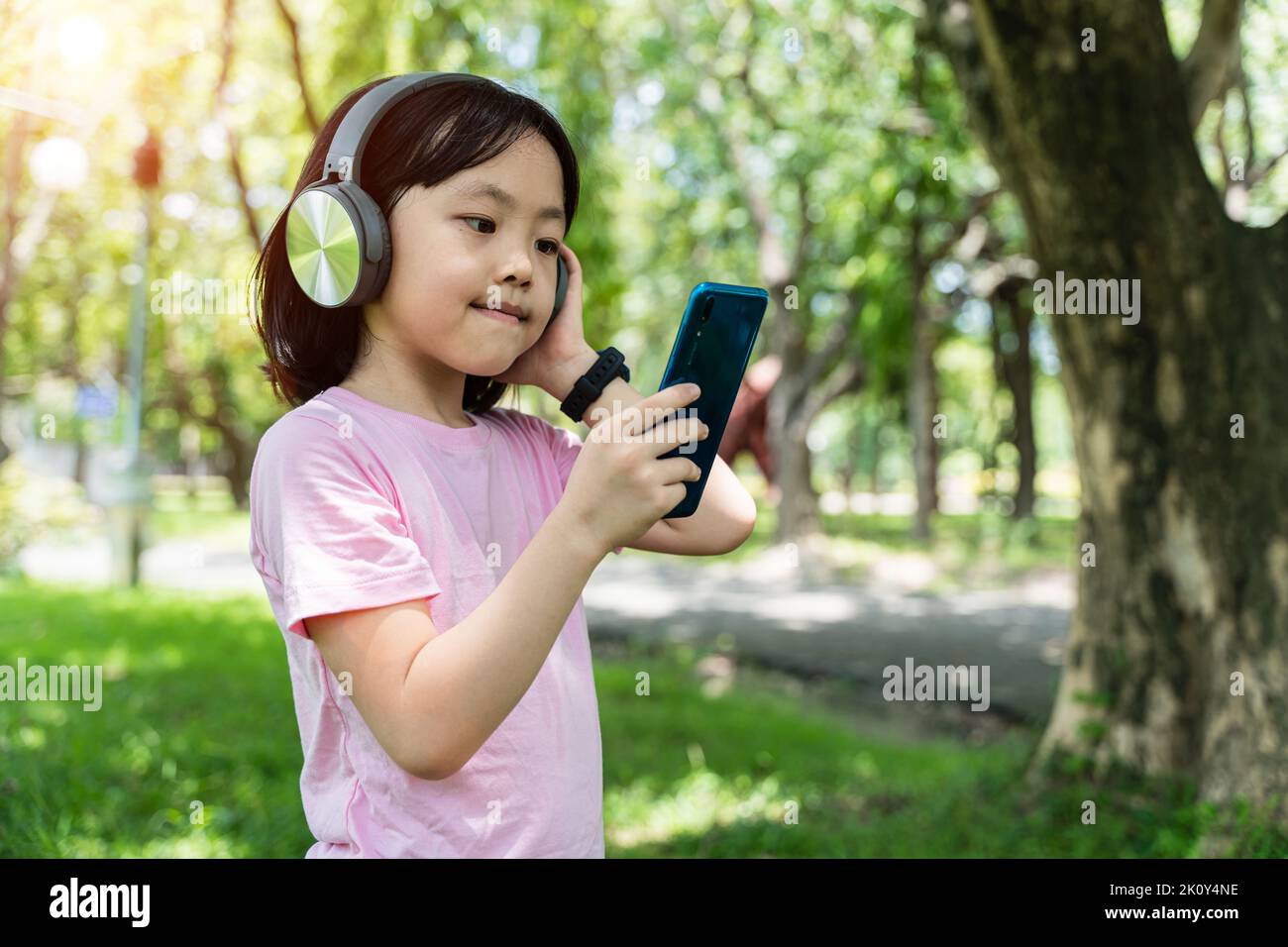 Primo piano della bambina asiatica con smartphone e ascolta la musica con le moderne cuffie wireless in un parco all'aperto. Foto Stock