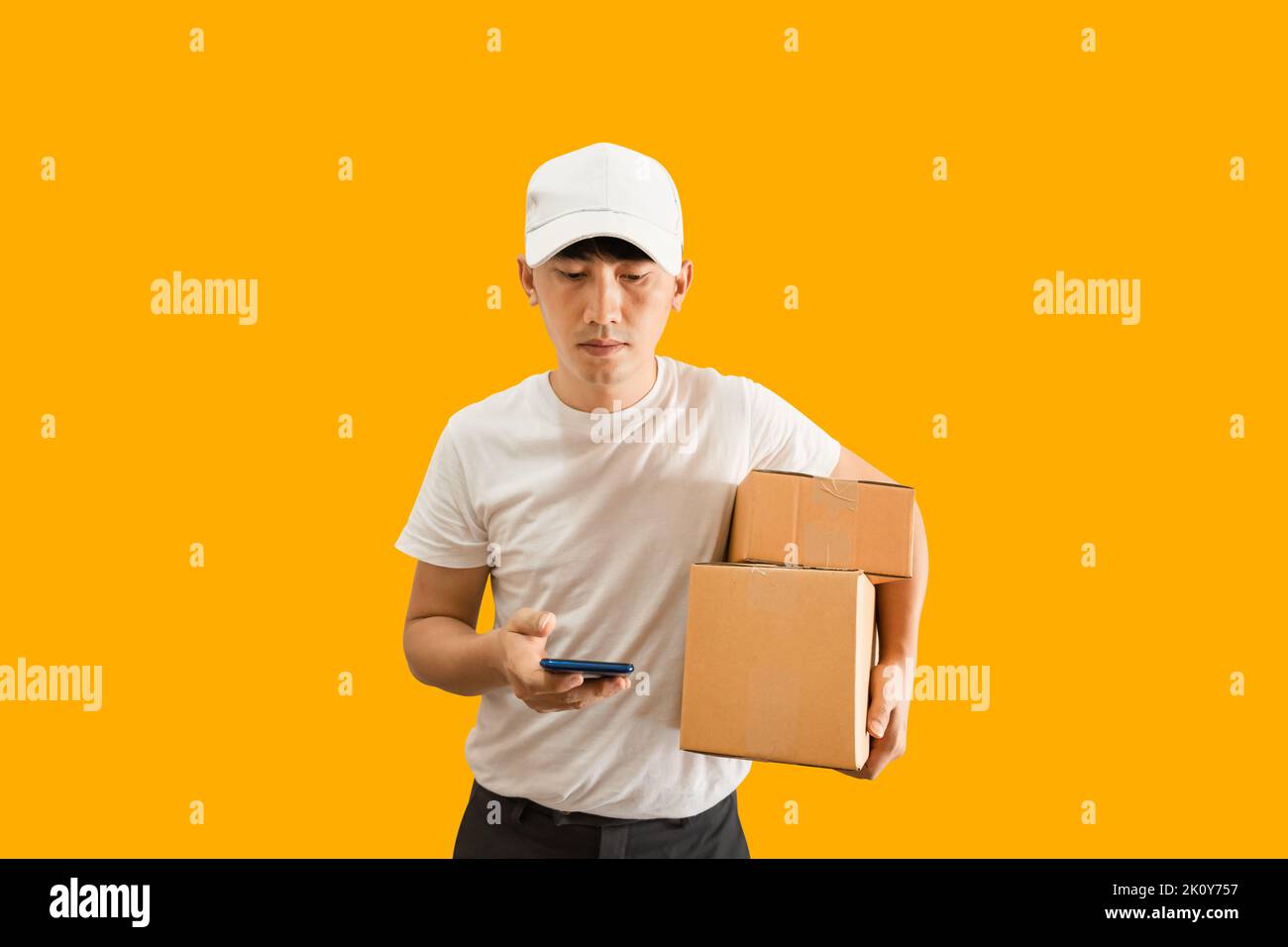 Giovane uomo asiatico con berretto e T-shirt bianca che tiene lo smartphone e la cassetta postale isolata su sfondo giallo. consegna rapida s Foto Stock