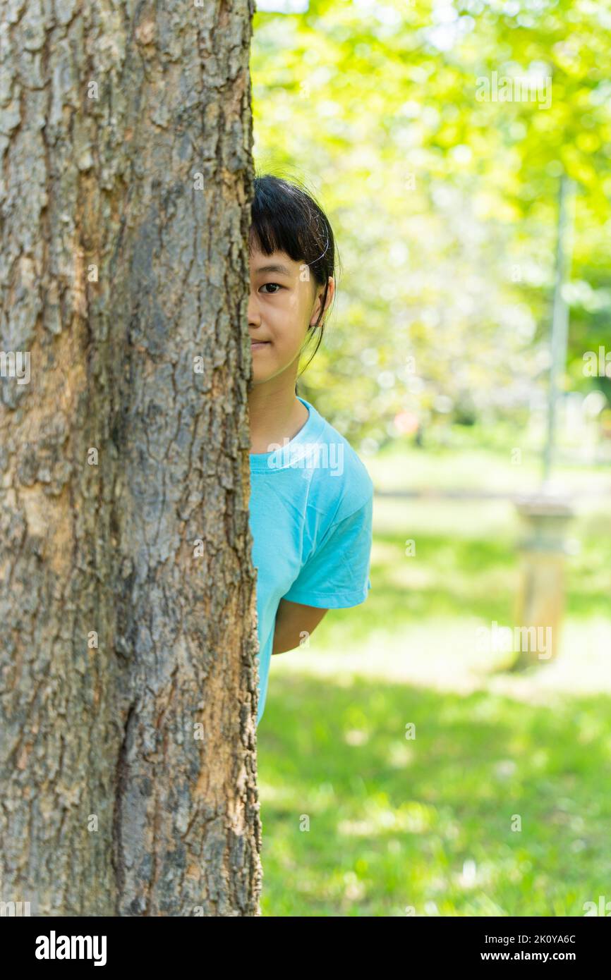 Bella bambina che sbircia fuori da dietro un albero. Ritratto di bambina che si gode la giornata estiva nel parco verde. Foto Stock