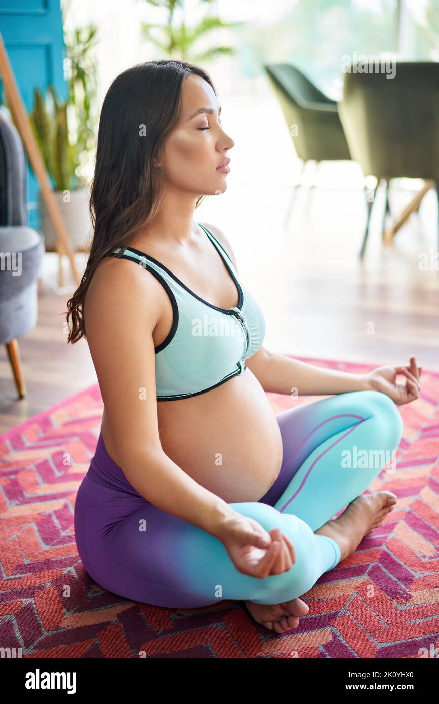 Donna sportiva incinta in abbigliamento sportivo pratica yoga, meditando in Lotus posa Padmasana a casa. Maternità cosciente. Stile di vita sano e cura del bambino Foto Stock
