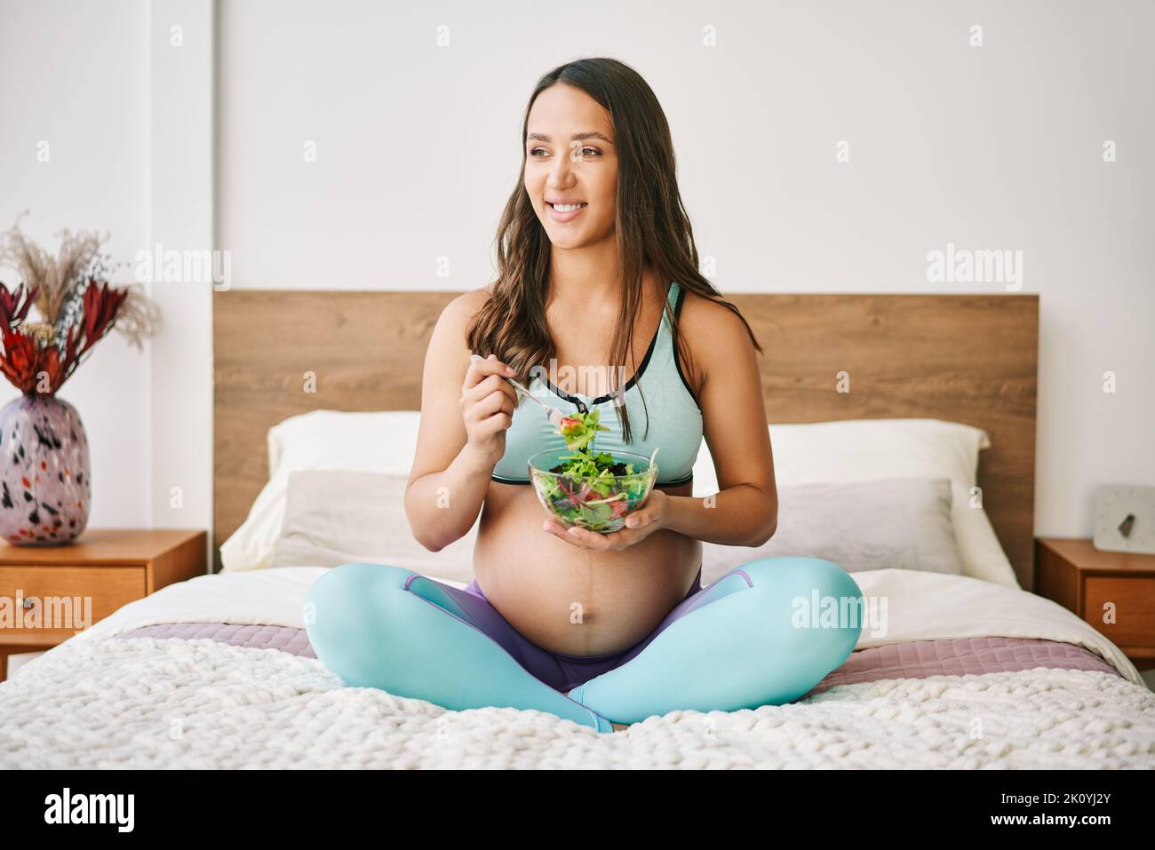 Ritratto di giovane donna incinta in abbigliamento sportivo mangiare un'insalata fresca seduta a letto. Maternità cosciente. Concetto di cibo sano Foto Stock