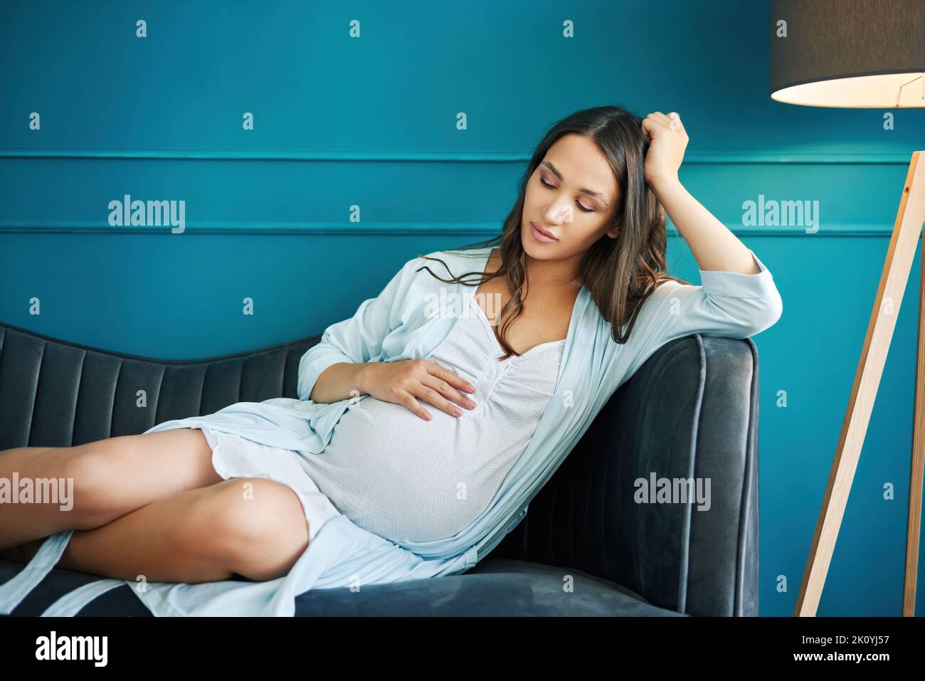 Ritratto di giovane donna incinta che tocca il ventre rilassarsi sul divano a casa moderna. Gravidanza, maternità, persone e concetto di aspettativa Foto Stock