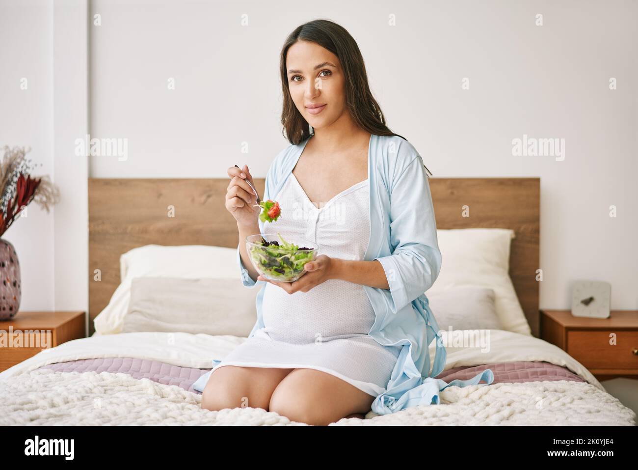 Ritratto di giovane donna incinta in pajama mangiare un'insalata fresca seduta a letto. Maternità cosciente. Concetto di cibo sano Foto Stock