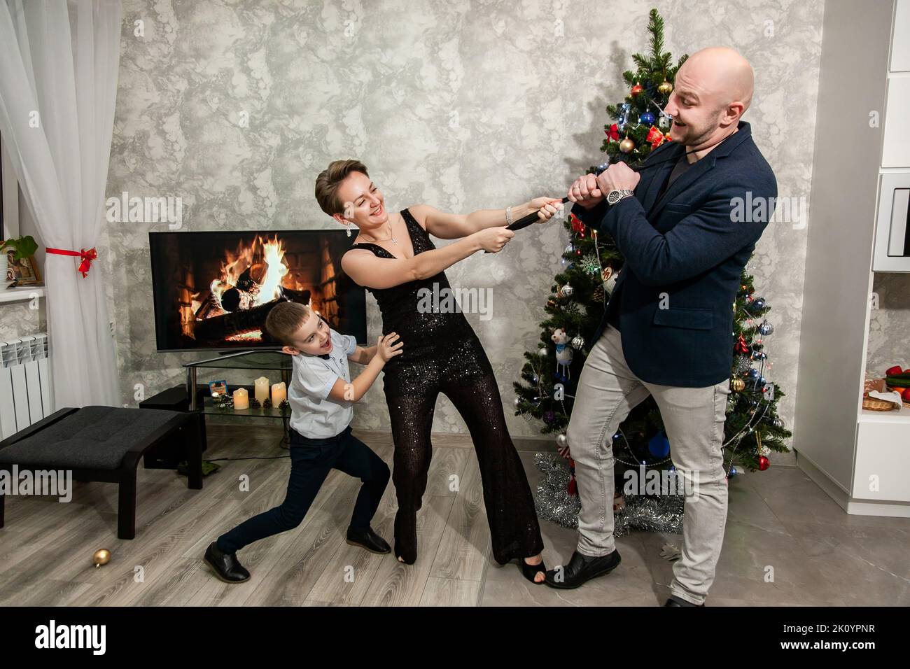 Famiglia mamma, papà e figlio pazzo intorno e giocare vicino all'albero di Natale nel loro appartamento. Moglie tira la cravatta del marito, il ragazzo aiuta Foto Stock