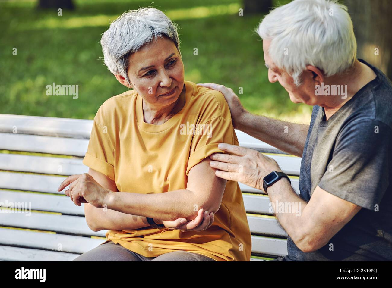Durante la passeggiata sportiva mattutina o facendo esercizi nel parco, la donna anziana di 60s anni si ferì la spalla o il braccio di presa del gomito seduto in panchina con disa premuroso Foto Stock