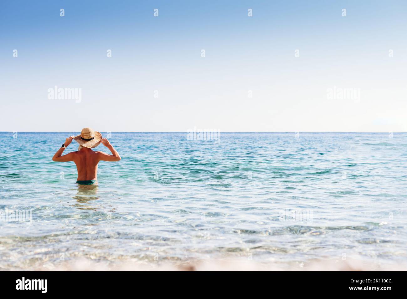 Bella donna bionda in un cappello di paglia che si erge sul mare su una spiaggia di Corfù Foto Stock