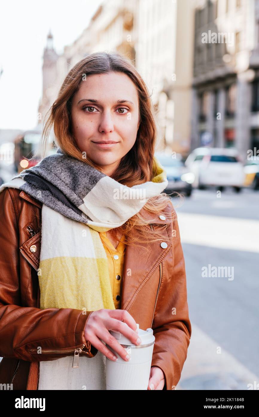 ritratto verticale di una giovane donna attraente pendolari in attesa in strada. Tiene un caffè take away e guarda la macchina fotografica Foto Stock