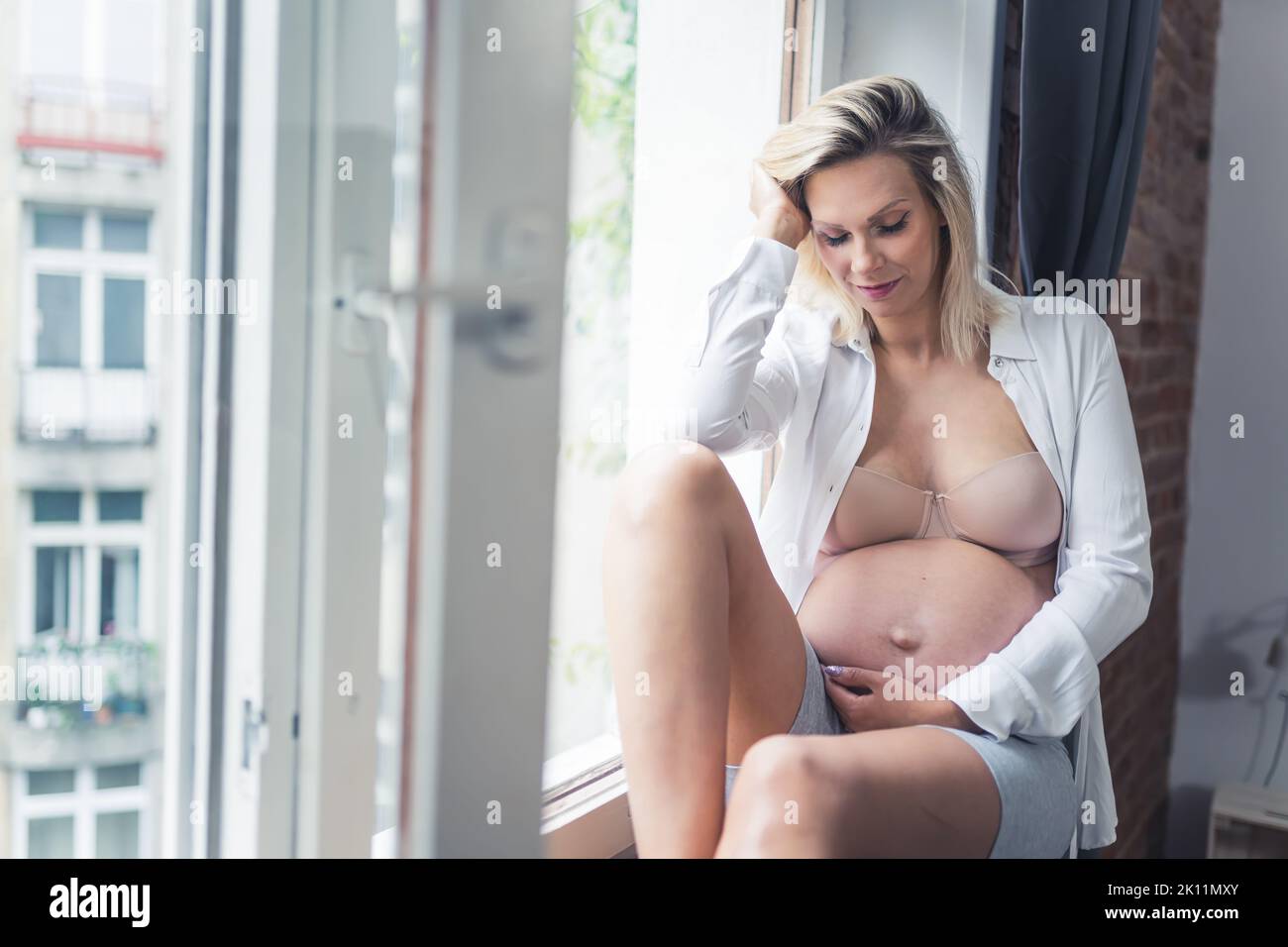 Donna sposata incinta seduta su un davanzale a casa, toccando il ventre e pensando di essere presto in grado di tenere il neonato. Foto di alta qualità Foto Stock