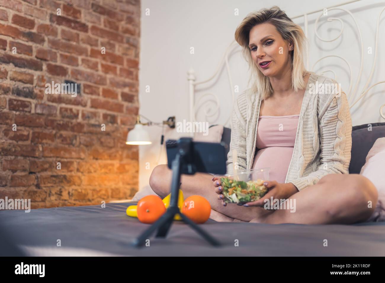 Moderna donna caucasica incinta seduta su un letto, mangiare un'insalata nutriente e snacking sulla frutta, parlando con i suoi amici in una videochiamata. Foto di alta qualità Foto Stock