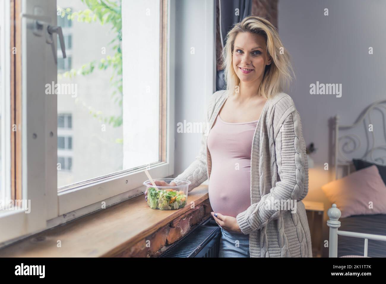 Mangiare sano durante la gravidanza. Giovane donna incinta caucasica in piedi vicino alla finestra con un nutriente, ricco di vitamine insalata pasto per la crescita di un bambino sano. Foto di alta qualità Foto Stock