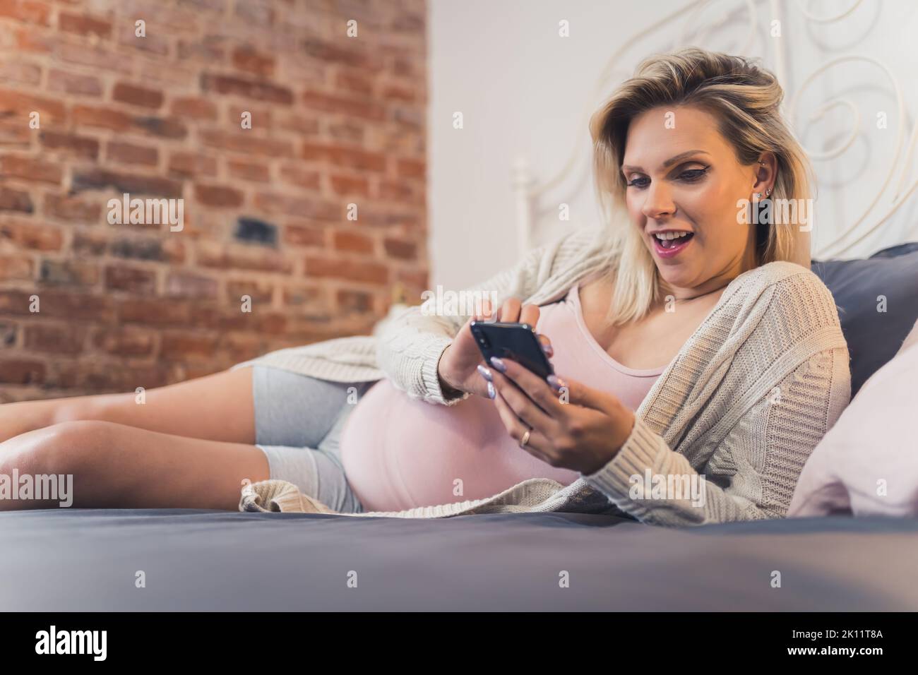 Giovane donna incinta che si rilassa a letto, guardando video divertenti sui social media tramite smartphone. Foto di alta qualità Foto Stock