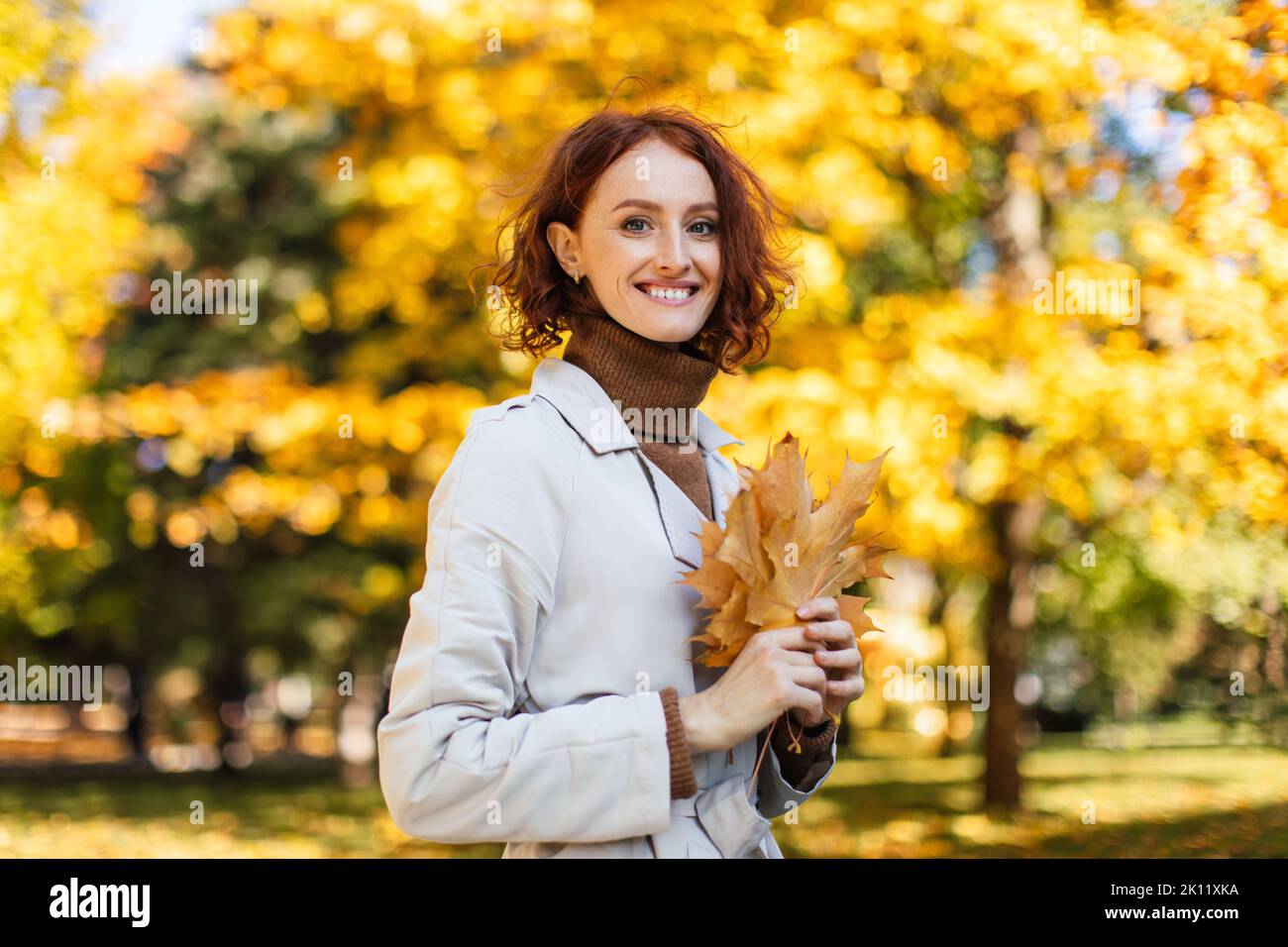 Camminare e stile di vita attivo. Felice donna caucasica millenaria rossa in impermeabile tenere foglie gialle Foto Stock