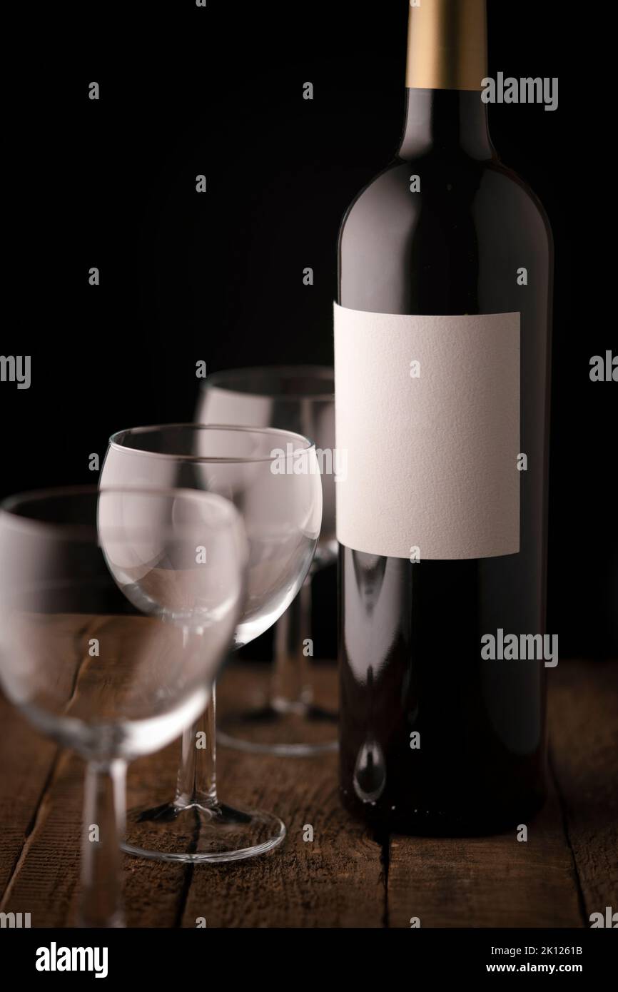 Una bottiglia di vino rosso con etichetta bianca vuota e tre piccoli bicchieri vuoti su un tavolo di legno Foto Stock