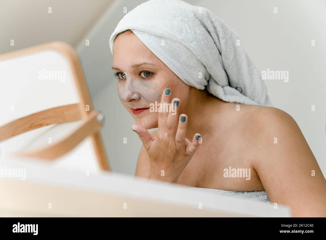 Giovane donna avvolta in asciugamano mette l'argilla cosmetica grigia sul suo viso mentre guarda in specchio. Primo piano Foto Stock