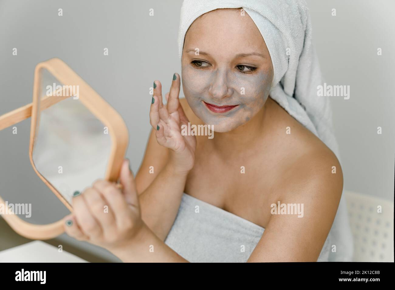La giovane donna mette l'argilla cosmetica grigia sul suo viso mentre guarda nello specchio, i suoi capelli e il suo corpo sono avvolti in asciugamano. Primo piano Foto Stock