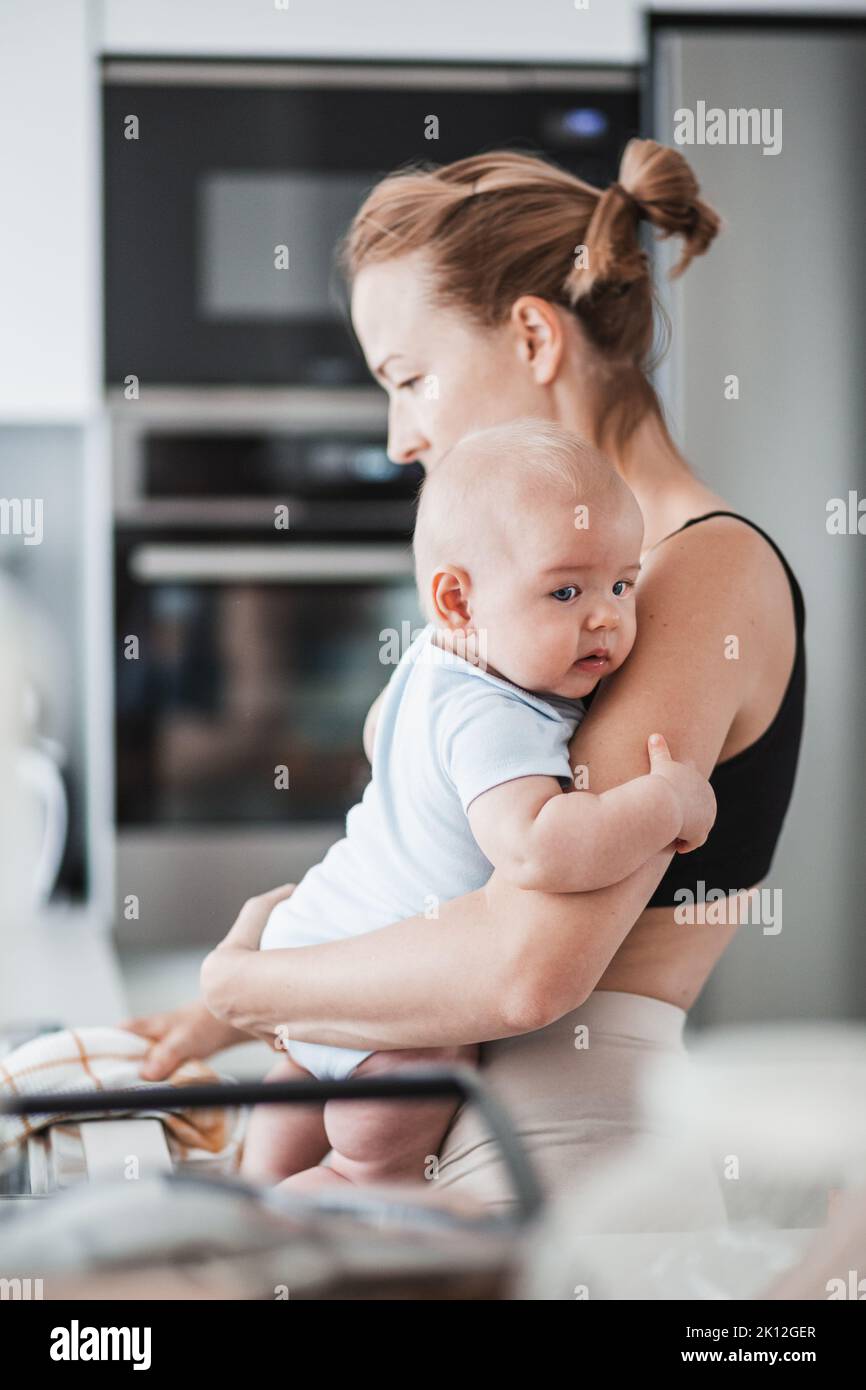 Donna che puliva il lavello della cucina con un panno dopo aver finito di lavare i piatti mentre tiene in mano il bambino di quattro mesi Foto Stock