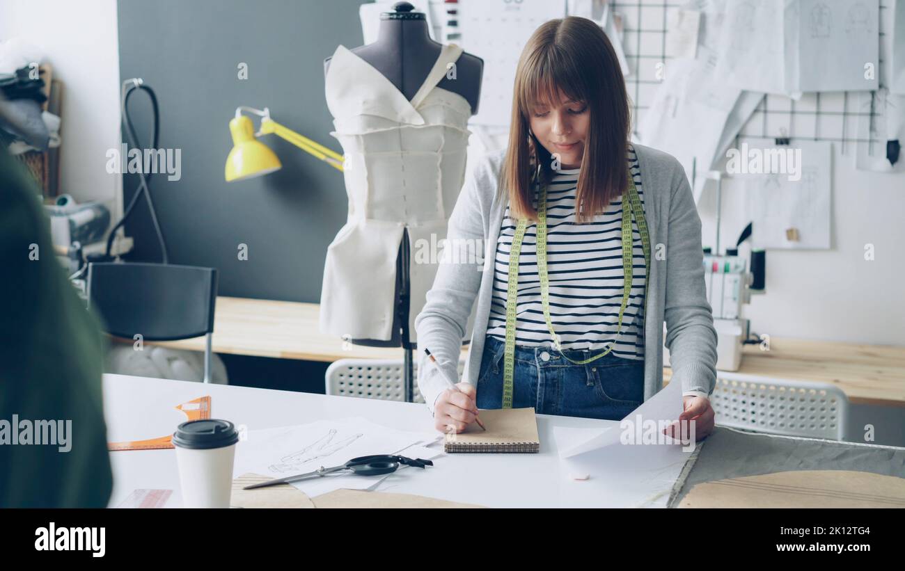 Giovane donna bella sta disegnando in blocco note guardando il disegno di abbigliamento mentre lavora nel suo studio di sartoria. Disegni appesi alla parete, oggetti da cucire luminosi sono visibili. Foto Stock