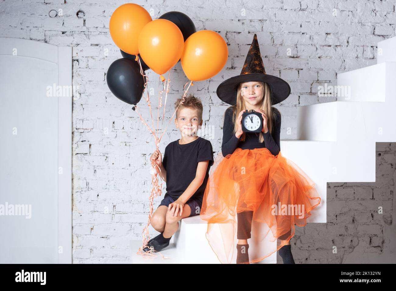 Halloween. I bambini in costumi e cappelli di strega creepy stanno tenendo una sveglia nera e palloncini neri e arancioni mentre si siedono sulle scale. Foto Stock