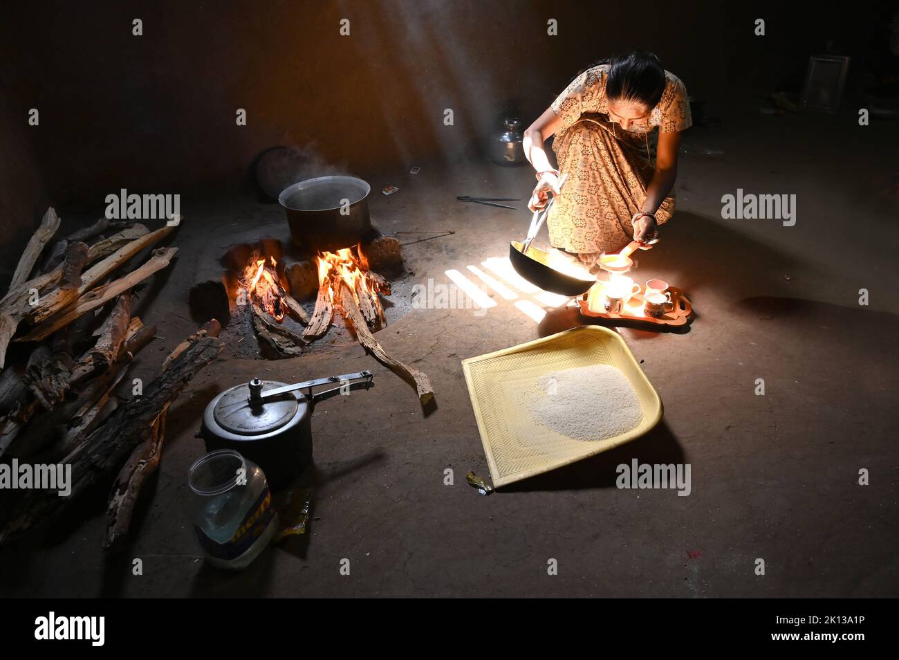 Luce del sole che efflue nella cucina tradizionale con pareti di fango con fuoco di legno, donna che fa chai, setaccio di riso di plastica, Kadana, Gujarat, India, Asia Foto Stock