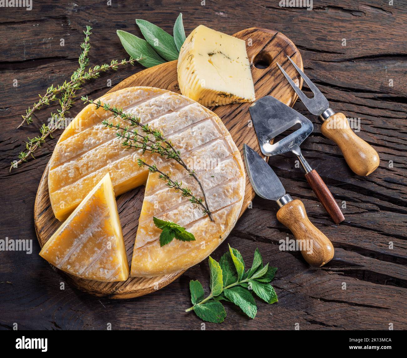 Testa di formaggio di pecora con erbe fresche e coltelli di formaggio su sfondo di legno. Foto Stock