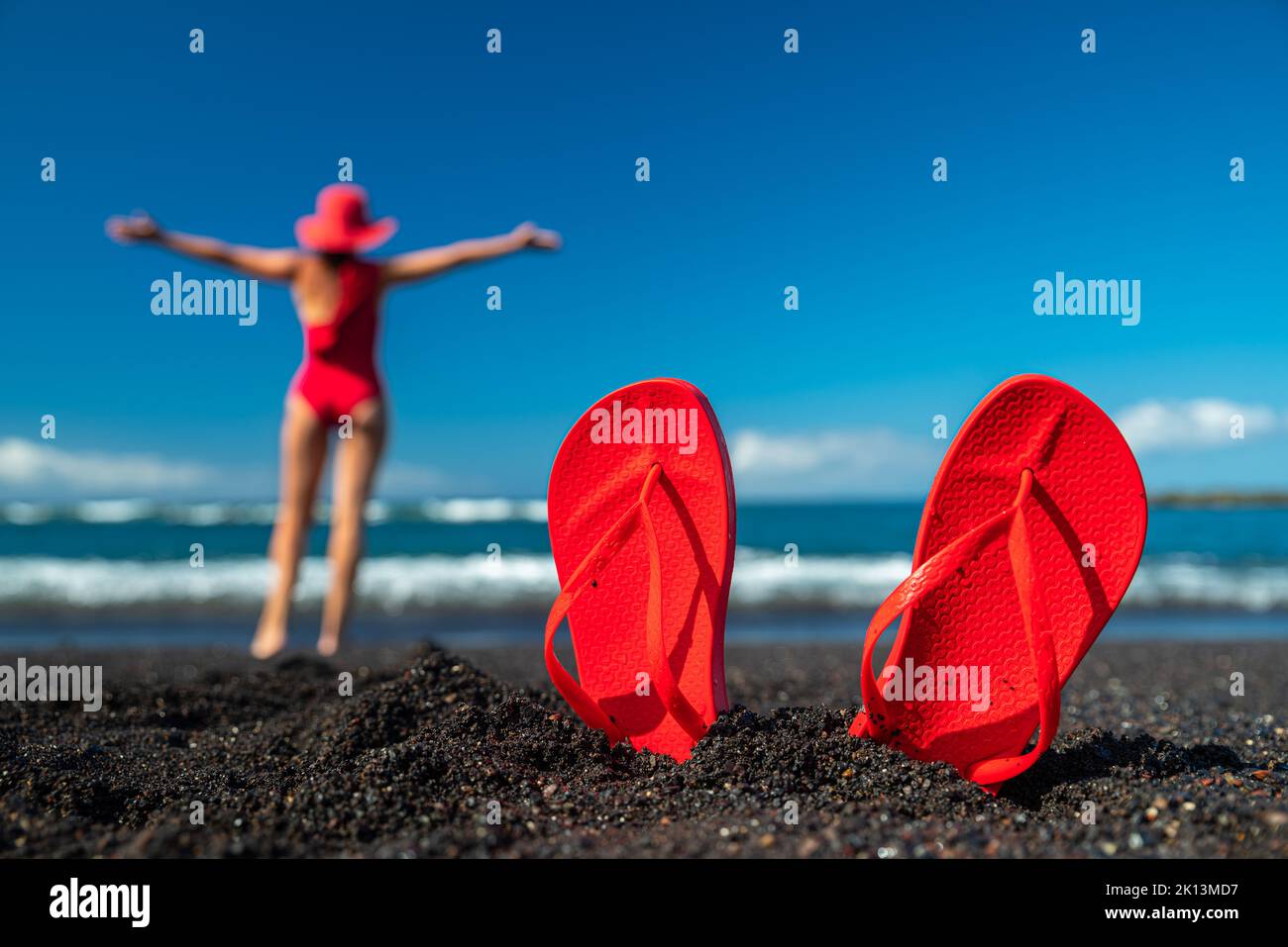 Infradito rosso nella sabbia nera e silhouette di donna in costume da bagno rosso in piedi sulla spiaggia dell'oceano. Concetto estivo. Foto Stock
