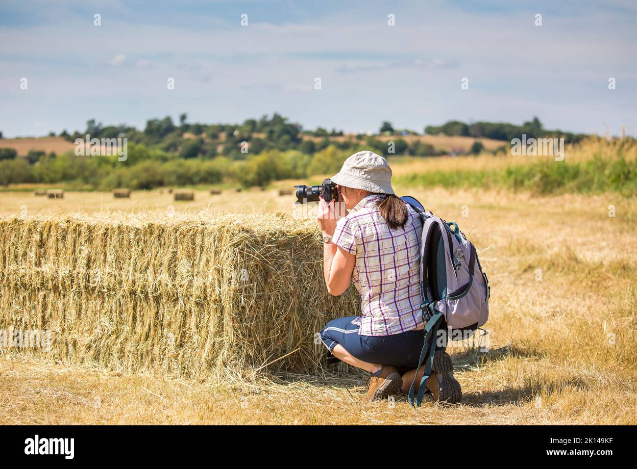Vista posteriore di una fotografa donna che si inginocchia da una balla di fieno scattando fotografie. Il tempo è caldo e soleggiato con una nuvola. Foto Stock