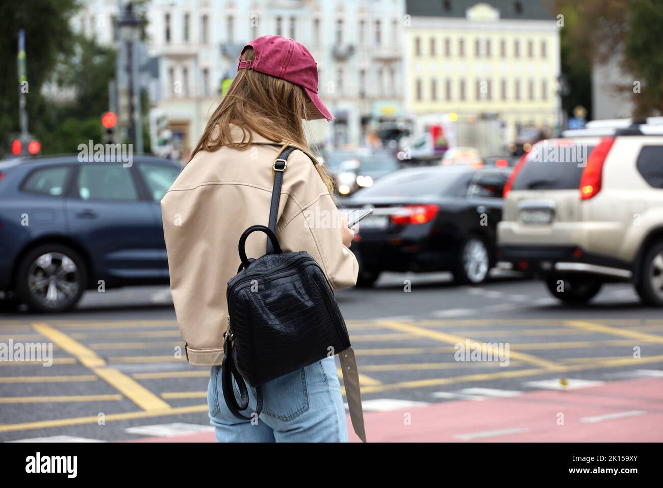 Ragazza che indossa jeans e giacca in pelle leggera in piedi sulla strada della città con smartphone in mani sullo sfondo delle auto Foto Stock