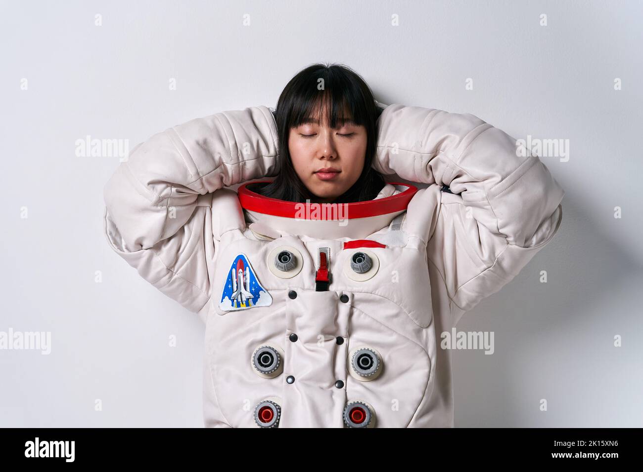 Giovane donna asiatica nella tuta spaziale che copre le orecchie e chiude gli occhi mentre cerca di prevenire disturbi contro sfondo grigio Foto Stock